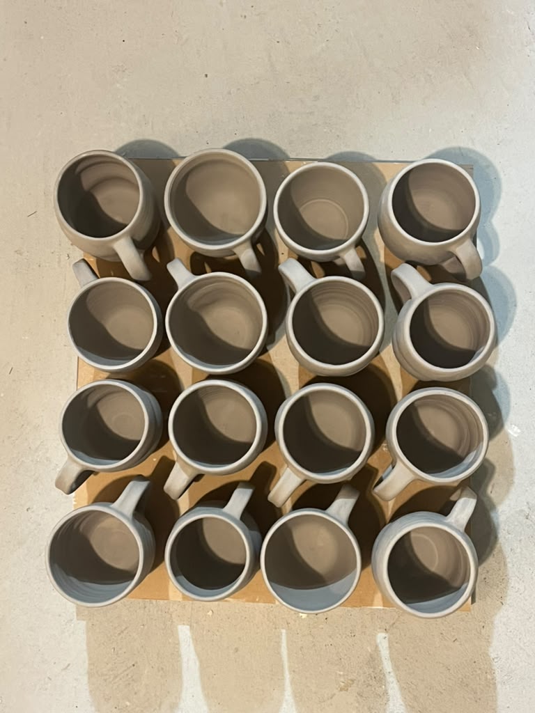Sixteen unglazed mugs with pulled handles, arranged in a grid on a board, seen from above.