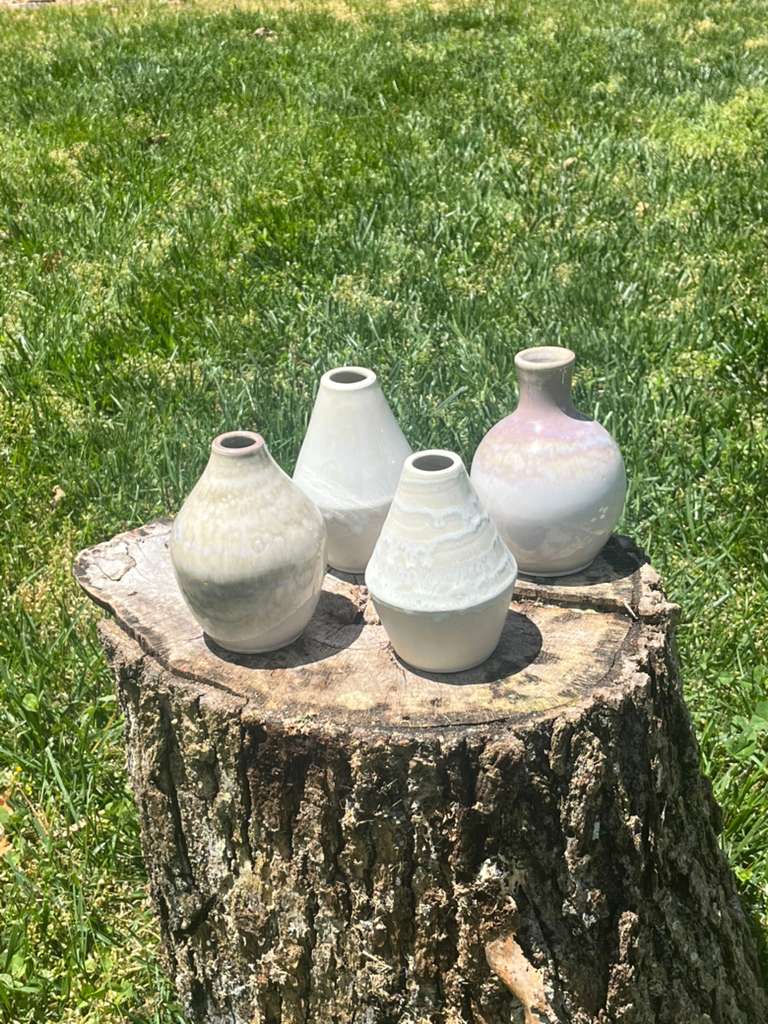 Four small bud vases with narrow necks in soft white, celadon and pale pink glazes, arranged on a tree stump.