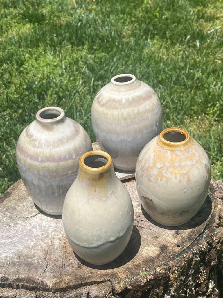 Four rounded stoneware vessels with pearly cream and lavender glazes and amber rims, on a tree stump in grass.