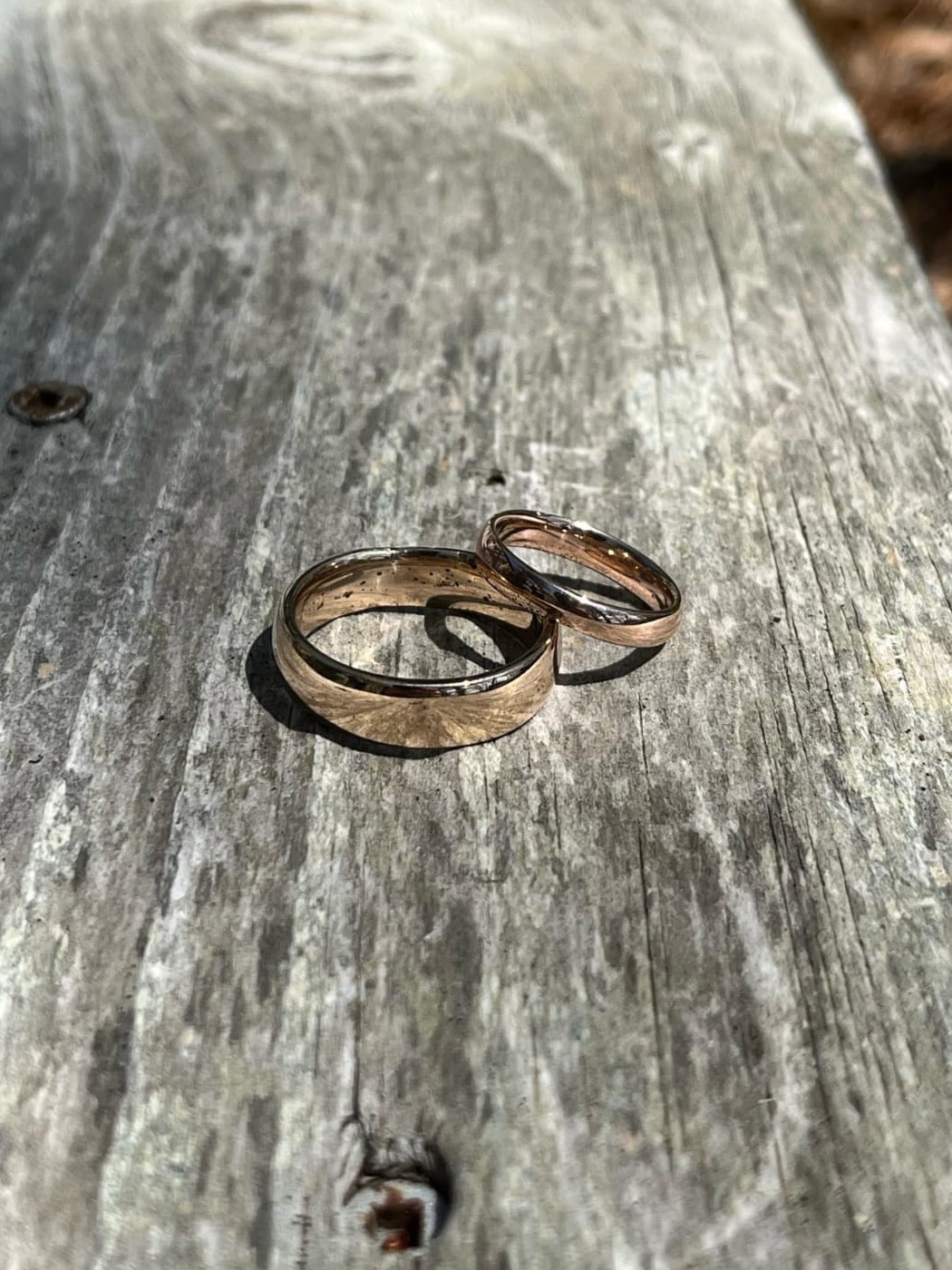Two warm gold-toned wedding bands, one wide and one slim, resting on a weathered grey board in sunlight.